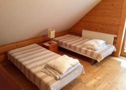 Dwa lóżka i szafka nocna, na niej lampka, ściany i podłoga drewniane, sufit biały skos