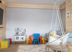 Podwieszany, sznurkowy fotel, szara kanapa, kącik z kolorowymi mebelkami dla dzieci oraz dziecięca kuchenka – zabawka, pod pomalowaną na szaro ścianą trzy pojemniki na zabawki