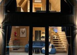Dwupiętrowy domek typu A-frame nocą. Przez wielkie przeszklone ściany widać oświetlone, drewniane wnętrze: na dole salon z niebieską kanapą i schodami, na górze sypialnię. Przed domkiem leży śnieg z widocznymi śladami stóp.