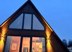 Drewniany domek typu A-frame o zmierzchu z widocznym na niebie księżycem. Przez duże przeszklone ściany widać ciepłe, oświetlone wnętrze salonu ze schodami. Zewnętrzne kinkiety oświetlają elewację budynku.