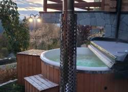 Drewniana strefa wypoczynkowa na świeżym powietrzu z jacuzzi, schodkami i ozdobnym metalowym słupem. W tle roślinność, oświetlenie ogrodowe oraz górski krajobraz o zachodzie słońca.