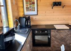 Drewniany aneks kuchenny z ciemnym blatem i szafkami w geometryczny wzór. Na blacie stoją mikrofalówka, ekspres do kawy i deska do krojenia, a w zabudowie widać piekarnik. Na ścianie wisi plakat oraz półka z kubkami.