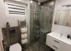 Łazienka w bieli i brązach, szklana kabina prysznicowa, biały sedes i szafka przy umywalce, nad nią lustro