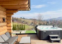 Drewniany taras przy domku z bali. Na tarasie znajduje się szara sofa ze stolikiem kawowym oraz jacuzzi ze schodkami. W tle roztacza się widok na małą miejscowość i zielone wzgórza pod błękitnym niebem z lekkimi chmurami.