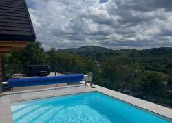 Prostokątny basen zewnętrzny z jasnoniebieską wodą na jasnym tarasie. Obok znajduje się zwinęta niebieska roleta basenowa oraz jacuzzi. W tle widoczne są zielone wzgórza, drzewa i domy pod zachmurzonym niebem.