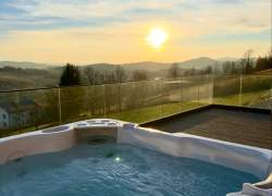 Białe jacuzzi z niebieską wodą na drewnianym tarasie ze szklaną balustradą. W tle widoczny panoramiczny krajobraz górski i zachodzące słońce na pogodnym niebie. Wokół rozciągają się zielone wzgórza i lasy.