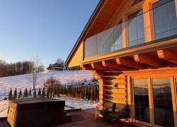 Drewniany domek z bali o zachodzie słońca. Na tarasie znajduje się czarne jacuzzi i fotele. W tle ośnieżone wzgórze z drzewami oraz błękitne niebo. Ciepłe światło słoneczne oświetla elewację budynku i szklaną balustradę balkonu.