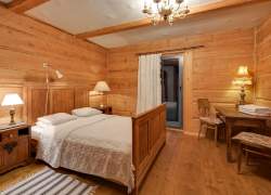Przestronna sypialnia, drewniane ściany i podłoga; małżeńskie łóżko z jasną pościelą, drewniana komoda, a na niej lampa, stół drewniany z dwoma krzesłami