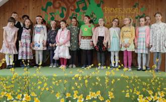 Uczniowie z klas 2 i 3 ustawieni na scenie w dwóch rzędach w kolorowych, wiosennych strojach; dzieci śpiewają.