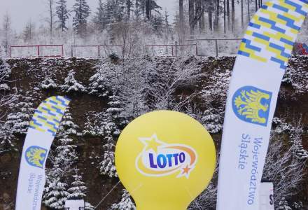 Żółty balon z napisem Lotto, strefa startu biegu narciarskiego z dwiema chorągiewkami  Województwa Śląskiego, w tle zimowa sceneria ze świerkami i porośnięte zbocze.