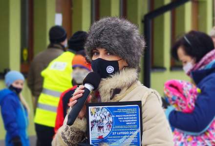 Ubrana w kożuszek i futrzaną czapkę konferansjerka biegu narciarskiego Aneta Legierska z mikrofonem w ręku, w tle dwoje dzieci i trzy osoby dorosłe -  uczestnicy wydarzenia
