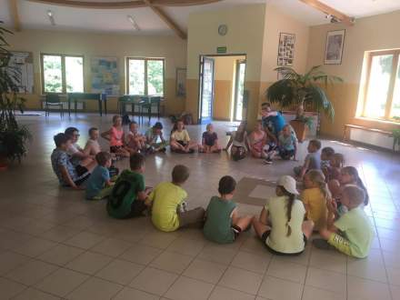 Dzieci siedzące na podłodze w kółeczku wraz z instruktorem; wnętrze to szkolny hol
