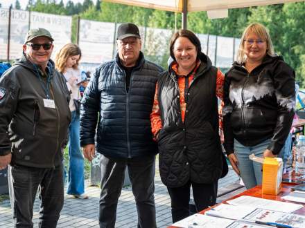 Cztery uśmiechnięte osoby – dwóch mężczyzn i dwie kobiety – pozują do zdjęcia przy stoisku organizacyjnym pod namiotem. Na stole przed nimi leżą dyplomy oraz dokumenty, a w tle widać otwartą przestrzeń.