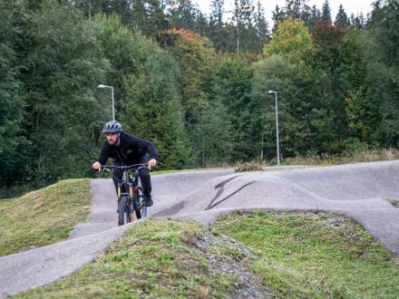 Mężczyzna w czarnym stroju i kasku dynamicznie jedzie na rowerze górskim po profilowanym torze pumptrack. Tor otaczają trawiaste pagórki, a w tle widać gęsty, wysoki las.