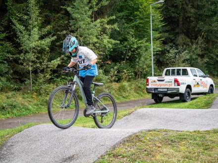 Młody rowerzysta w kasku i niebiesko-białej koszulce z numerem startowym jedzie po torze pumptrack. W tle stoi biały pick-up z napisem zlotygron.pl oraz gęsty, zielony las.