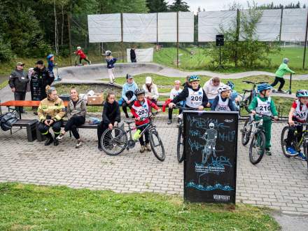 Widok na plenerowy tor pumptrack z uczestnikami zawodów rowerowych. Dzieci w kaskach stoją z rowerami, a na ławkach i obok toru siedzą widownia oraz organizatorzy. W tle widać ogrodzenie oraz drzewa.