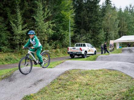 Chłopiec w kasku i zielonym dresie jedzie na rowerze górskim po torze pumptrack. W tle stoi biały pick-up z napisem zlotygron.pl oraz namiot. Całość otacza gęsty, zielony las.