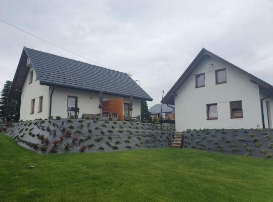 Widok zewnętrzny domków - białe murowane obiekty z szarymi dachami, dookoła zieleń trawy i nasadzenia krzewów ozdobnych
