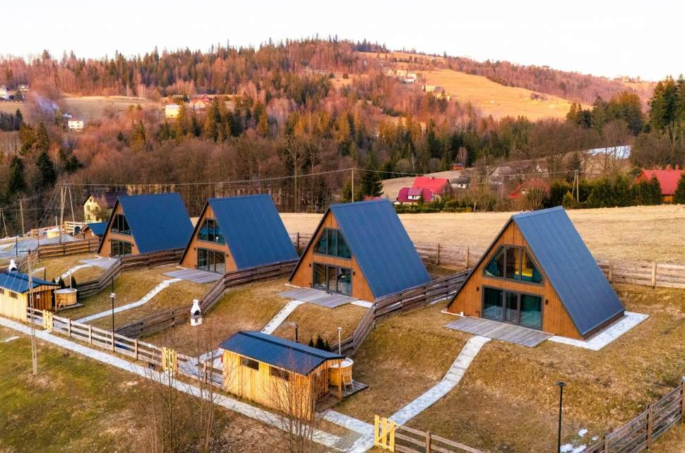 Widok z góry na cztery drewniane domki typu A-frame z ciemnymi dachami, ustawione w rzędzie na ogrodzonym zboczu. W tle widać malownicze, zalesione wzgórze w jesiennych barwach pod błękitnym niebem z chmurami.