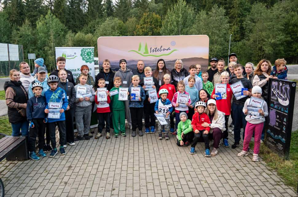Grupowe zdjęcie pamiątkowe dzieci i dorosłych na świeżym powietrzu. Dzieci trzymają dyplomy i spoglądają w stronę obiektywu. W tle widnieje zielona ścianka reklamowa z napisem Istebna oraz malowniczy, leśny krajobraz.