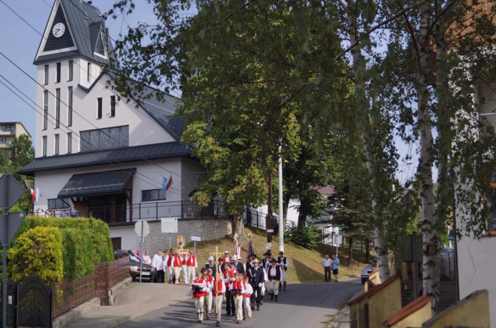 Uroczysta procesja idąca ulicą. Uczestnicy, w tym osoby w regionalnych strojach góralskich i mundurach, niosą krzyż oraz poczty sztandarowe. W tle stoi nowoczesny kościół z wieżą zegarową, otoczony bujnymi zielonymi drzewami.